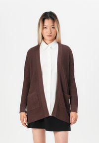 Brown ribbed cardigan with open front, two front pockets, and long sleeves, worn over a white button-up shirt and black skirt.