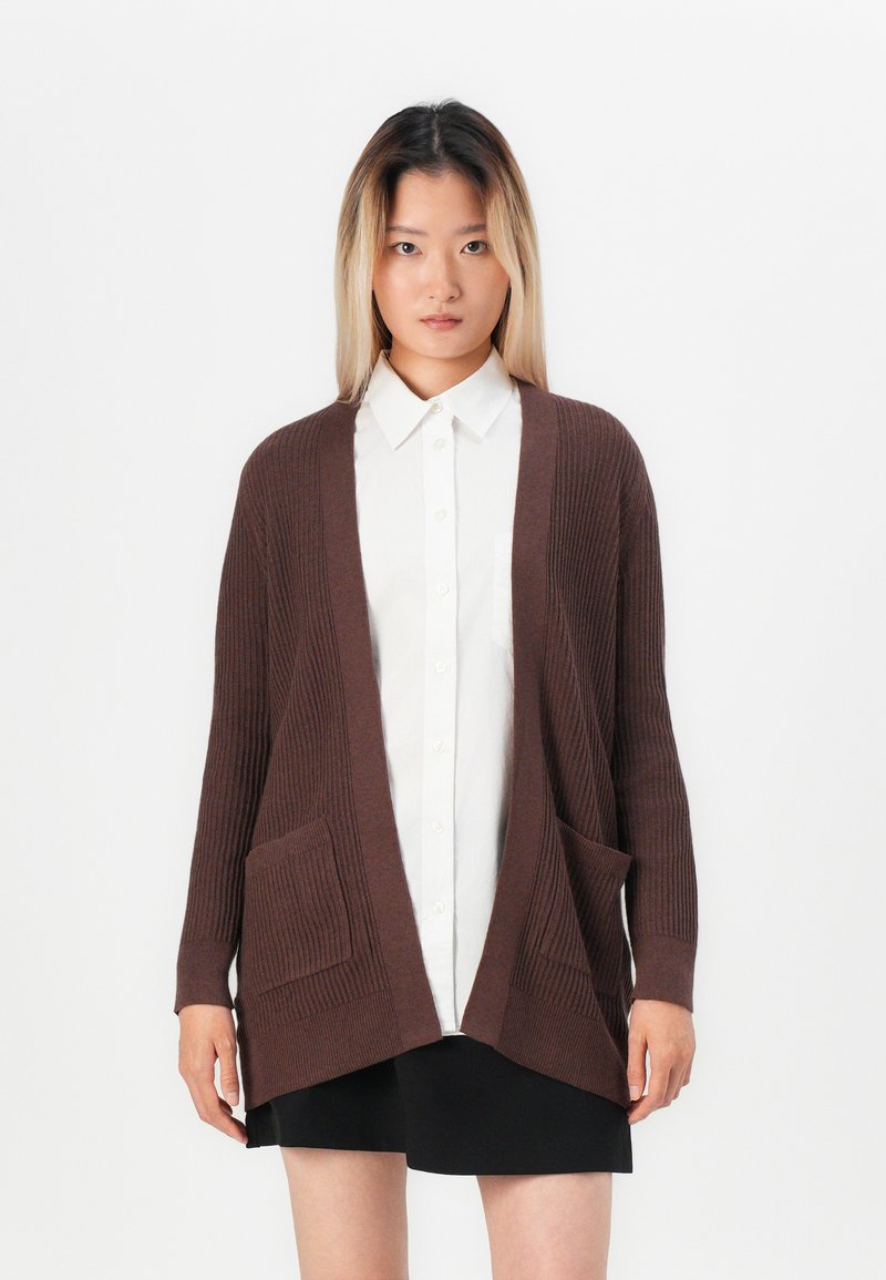 Brown ribbed cardigan with open front, two front pockets, and long sleeves, worn over a white button-up shirt and black skirt.