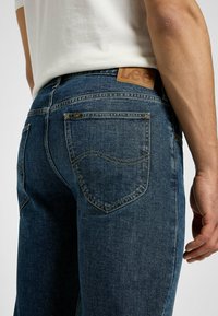 Los jeans de mezclilla azul oscuro presentan una etiqueta de cuero con "Lee", costuras visibles y un diseño clásico de cinco bolsillos con bolsillos traseros curvados.