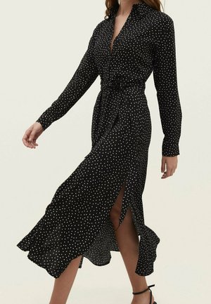Robe midi noire à manches longues à pois blancs, boutonnée à l'avant, ceinture à la taille, fente sur le côté, portée par une femme marchant en sandales à talons.