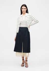 Γυναίκα που φοράει λευκό ριγέ ζακέτα και τοπ, μπλε σκουρόχρωμη φούστα με σχίσιμο μπροστά, φόρεμα με κρεμ δαντέλα από κάτω και μαύρα ψηλοτάκουνα με λουράκια.