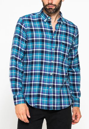 Uomo che indossa una camicia a quadri blu, viola, bianca e nera con bottoni e maniche lunghe e pantaloni neri, su sfondo bianco.