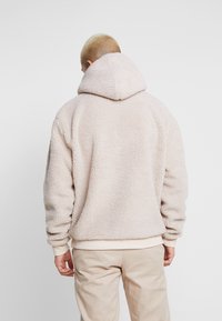 Beige Fleece-Hoodie mit einer strukturierten, plüschigen Oberfläche, Kängurutaschen und einer Kapuze. Bündchen und Saum aus glattem Material.