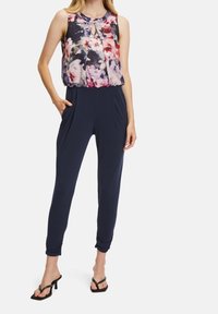 Femme portant un chemisier sans manches à motif floral avec encolure en trou de serrure, pantalon bleu marine fuselé avec poches latérales, et sandales noires à talons.