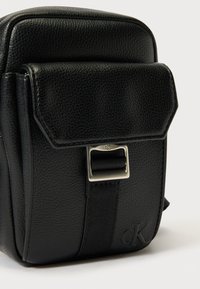 Mustast nahast kott tekstuurse pinnaga, ristkülikukujuline, klapiga sulgur ja hõbedased detailid, millel on „ck“ logo.