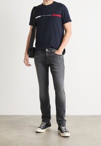 Navy T-shirt with horizontal red and white stripe, paired with dark gray jeans and black canvas sneakers, carrying a black messenger bag.