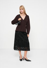 Donna che indossa un maglione a V con bottoni di color marrone scuro, una gonna midi nera con texture, e tacchi a slingback neri, mentre tiene una piccola borsa nera.