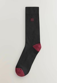 Musta sukka, jossa on yhtenäinen muotoilu ja viininpunainen kärki sekä kanta. Yläosassa on pieni punainen kirjailtu logo.