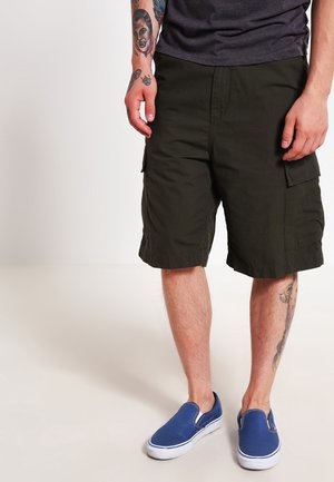 Homme portant un short cargo vert foncé, des chaussures bleu slip-on et un t-shirt gris foncé, avec des tatouages visibles sur les bras et les jambes.