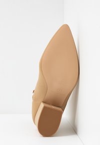 Chaussure beige à bout pointu avec un petit talon bloc, semelle tournée vers l'avant, positionnée contre des murs d'angle blancs.