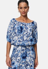 Blouse florale bleue avec manches bouffantes, décollet�é bardot et tissu texturé. Présente des motifs floraux complexes en bleu et blanc.
