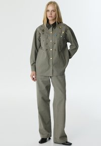 Khaki overhemd met knopen, versierd met pins en patches, in combinatie met bijpassende broek. Heeft een puntige kraag en twee zijzakken.
