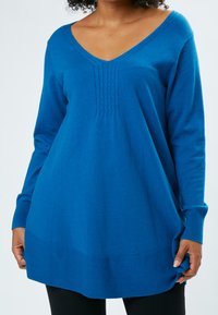 Blauer Strickpullover mit V-Ausschnitt, langen Ärmeln und vertikalen Rippendetails auf der Vorderseite. Weiche Textur und entspannte Passform, bis zur Mitte des Oberschenkels reichend.