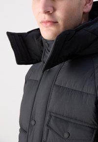 Zwarte, pufferjack met een hoge kraag, drukknoopjes en twee borstzakken. Glad, gestructureerd materiaal met een matte afwerking.