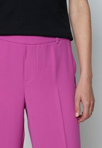 Un pantalon rose sur mesure avec un design à l'avant plat, présentant une texture lisse et une poche sur le côté. Associé à un haut noir, offrant une coupe décontractée.