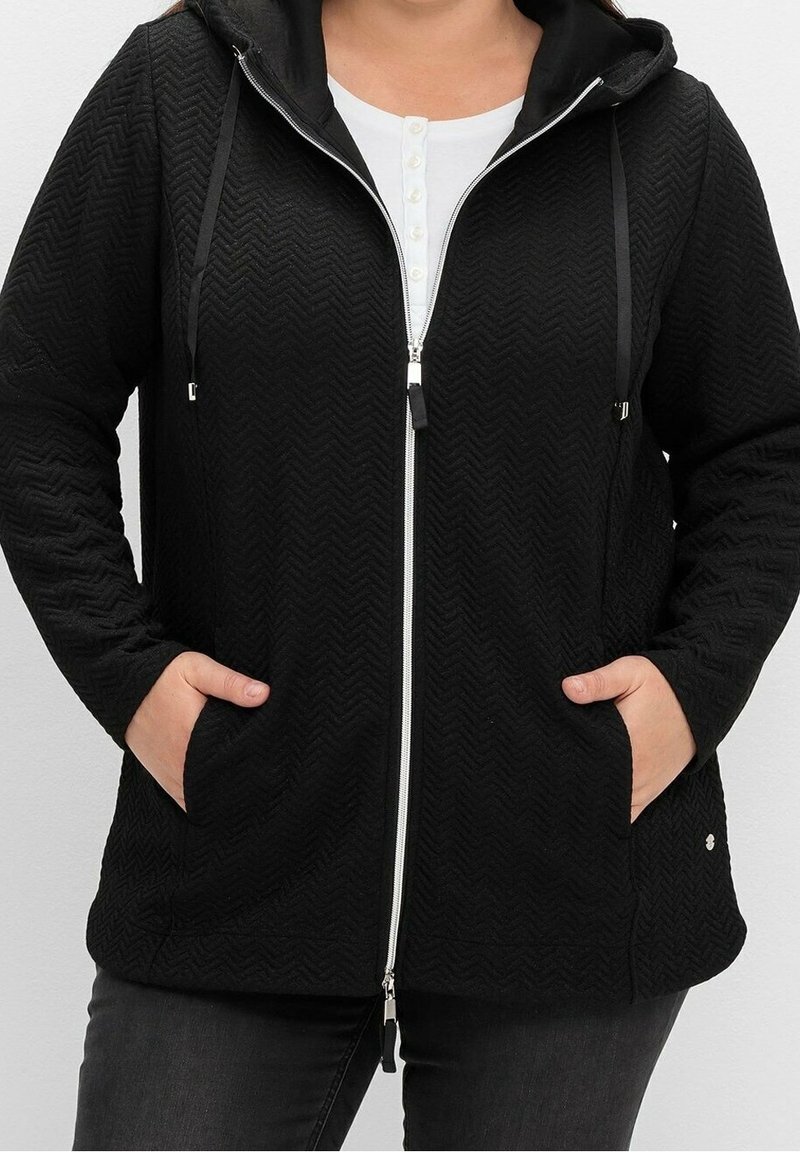Donna che indossa una felpa nera con zip e texture, dotata di cappuccio e tasche, abbinata a una camicia bianca con bottoni e pantaloni scuri.