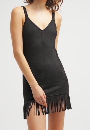 Vrouw die een korte zwarte jurk zonder mouwen draagt met dunne bandjes en franjes langs de zoom, staand tegen een effen achtergrond.