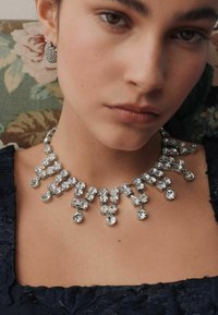 Silberne Halskette mit mehreren klaren Kristallen in unterschiedlichen Formen, die in einem geschichteten Design mit hängenden Akzenten angeordnet sind.