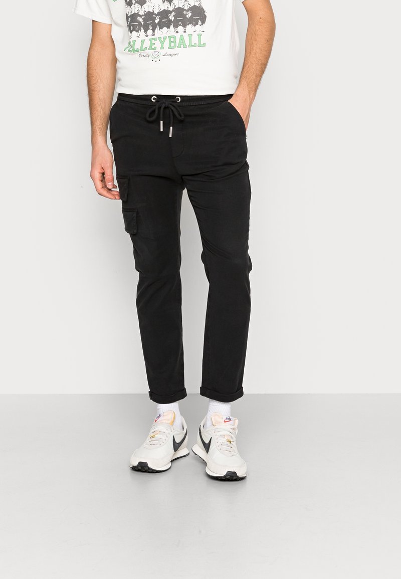 Černé cargo kalhoty s páskem na stažení v pase, se bočními kapsami a zúženým střihem. Nosené s bílými teniskami a tričkem s potiskem.