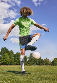 Puma grønn mønstret drakt, svarte shorts med blå detaljer, hvite sokker, svarte fotballsko, balanserer en hvit fotball på en fot.