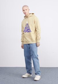 Sudadera marrón con logo morado, jeans azules y zapatillas blancas. La sudadera tiene un bolsillo canguro y puños acanalados. Diseño simple y casual.