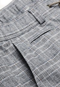 Gros plan sur un pantalon en tissu à carreaux gris montrant une passants de ceinture, la ceinture, une poche latérale et un motif de tissu à carreaux détaillé.