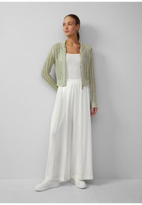 Jasnozielona, dziergana kurtka o skróconym fasonie z złotym zamkiem, zestawiona z białą bluzką i szerokimi białymi spodniami oraz białymi trampkami.