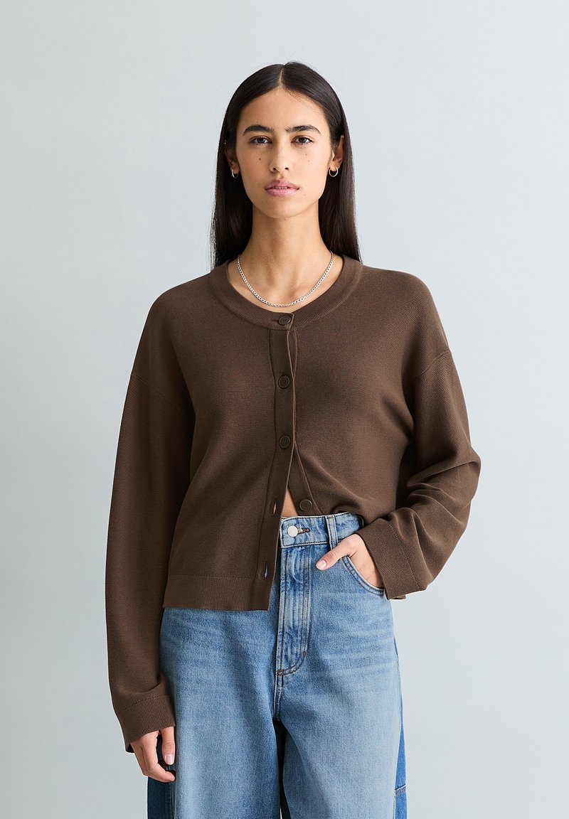 Femme aux longs cheveux bruns portant un cardigan marron boutonné et un jean taille haute bicolore bleu, debout avec une main dans la poche.