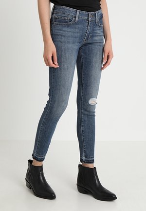 Jeans ajustados en denim azul oscuro con detalle de rodillas desgastadas. Cuenta con cinco bolsillos y un dobladillo deshilachado en el tobillo. Combinados con botas negras de tobillo.