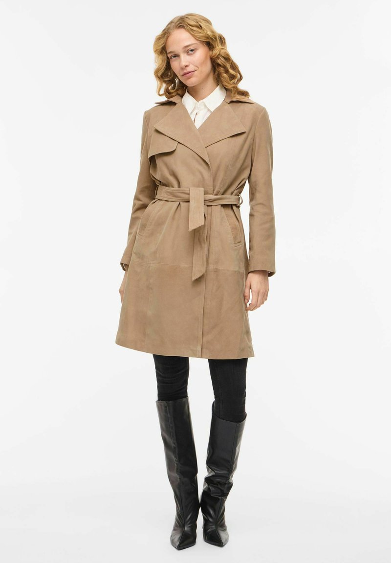 Žena s kučeravými blond vlasmi, oblečená v béžovom trenčkot s opaskom, bielej košeli, čiernych nohaviciach a čiernych čižmách nad kolená, stojaca na bielom pozadí.