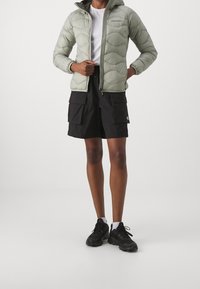 Ljusgrön pufferjacka med huva, med ett quiltat mönster, hel dragkedja och synliga fickor. I kombination med svarta cargoshorts och svarta sneakers.