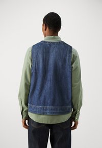 Denimvest i mørkeblå, båret over en lys grøn skjorte, med rygstykke og sidelommer, parret med mørke jeans.