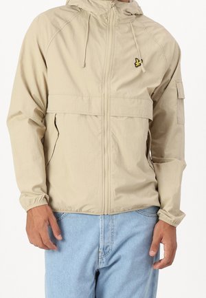 Osoba nosząca beżową kurtkę z kapturem, suwakiem z przodu, bocznymi kieszeniami i małym żółtym logo ptaka na piersi, zestawiona z jasnoniebieskimi jeansami.