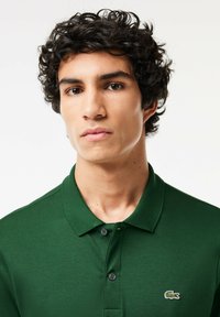 Groene polo-shirt van katoen met een klassieke kraag, twee zwarte knopen en een klein geborduurd krokodillogo op de borst.