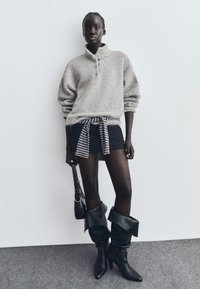 Sivi flis pulover s ovratnikom na zip, u kombinaciji s crnom mini suknjom koja ima prugasti pojas. Crne čizme do koljena s špicastim vrhom. Crna torba.
