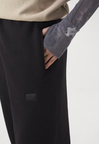 Hand in pocket van zwarte broek met logo, draagt een beige top en een grijze langemouwen shirt met een lichte vlek op de manchetten.