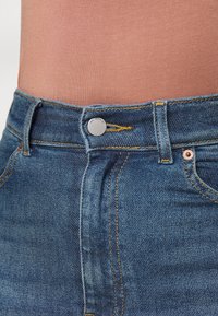 Närbild av midjan på blå denimjeans med metalldosa märkt "DR. DENIM" och gul sömnad, kombinerat med en ljusbrun topp.