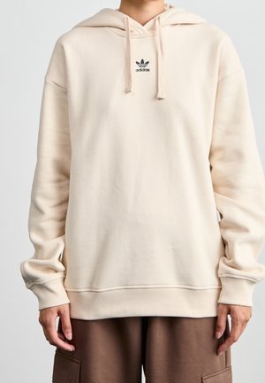 Osoba nosząca kremową bluzę z kapturem Adidas z logo na piersi oraz brązowe spodnie typu cargo, stojąca twarzą do przodu na prostym tle.