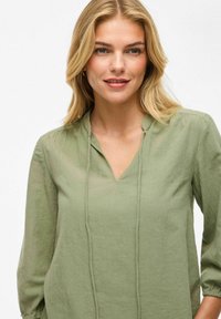 Blusa verde oliva realizzata in tessuto leggero, con scollatura a V, dettaglio con cordoncino e maniche a tre quarti con polsini arricciati.