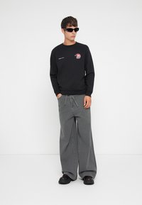 Fekete pulóver logóval, szürke bőszárú nadrág és fekete napszemüveg. Sima anyag, laza kényelem, és feltűnő varrási részletek.