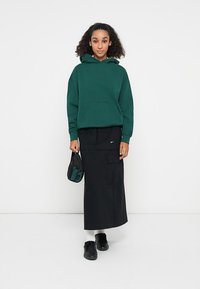 Sweatshirt verde com um bolso canguru, combinado com uma saia maxi preta com bolsos de carga. Ténis pretos e uma mala pequena completam o look.