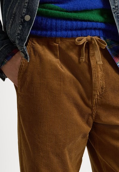 Pantaloni din catifea groasă maro, cu o textură ribbed, având talie cu șnur și buzunare laterale. Asortați cu un pulover albastru purtat sub o jachetă din denim.