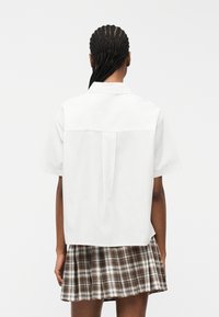 Άτομο με κοτσίδες που φοράει χαλαρό λευκό πουκάμισο με κοντά μανίκια και καφέ πλισέ φούστα με καρό σχέδιο, εμφανιζόμενο από πίσω.