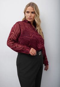 Genomskinlig vinröd blus med vågig mönstring, knäppning framtill, långa ärmar, i kombination med högmidjade svarta byxor och ett bälte.