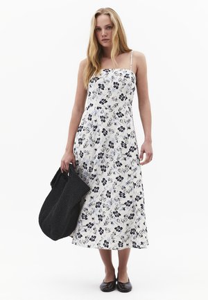 Vrouw in een witte midi-jurk met bloemenprint en dunne bandjes, houdt een grote zwarte geweven tas vast en draagt zwarte platte schoenen op een witte achtergrond.