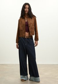 Frau mit langem dunklem Haar trägt eine braune Jacke mit Knöpfen, ein bauchfreies dunkelrotes Top, weit geschnittene dunkle Jeans mit hochgekrempelten Säumen und beige Schuhe.