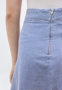 Close-up van een blauw-wit gestreepte rok met een zilveren ritssluiting aan de achterkant, gedragen door een persoon met een zichtbaar linkerarm en blote huid.