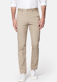 Bežové bavlněné kalhoty s rovný fit, obsahující boční kapsy, přední zapínání na knoflík a hladký povrch. Nosené s bílými teniskami.