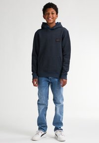 Felpa blu navy realizzata in tessuto testurizzato, con tasca a marsupio, polsini a coste e una patch con logo. Abbinata a jeans azzurri e sneakers bianche.