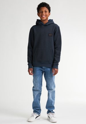 Sweat à capuche bleu marine en tissu texturé, avec poche kangourou, poignets côtelés et une patch logo. Assorti à un jean bleu clair et des baskets blanches.